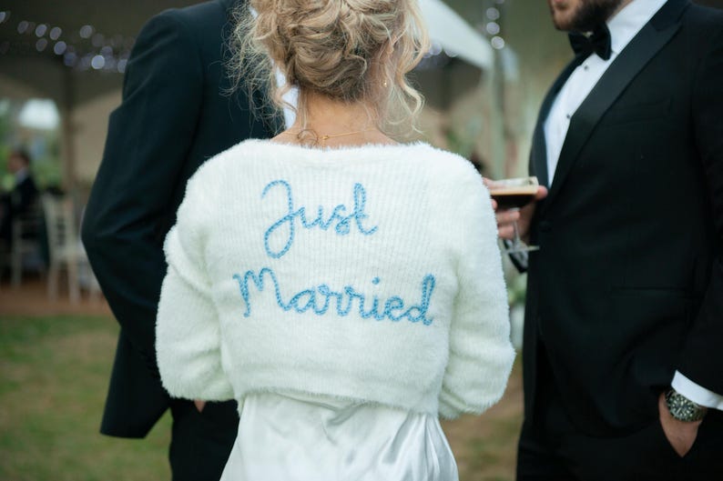 Puede incluir: Un bolero blanco y esponjoso con las palabras "Just Married" bordadas en azul claro. El su&eacute;ter se usa sobre un vestido blanco. La imagen est&aacute; tomada en una boda al aire libre.