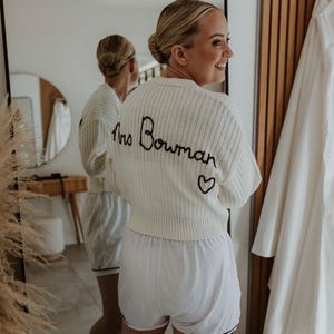 May include: A white knitted cardigan with the text "Mrs. Bowman" embroidered on the back. The cardigan has a small heart embroidered below the text.