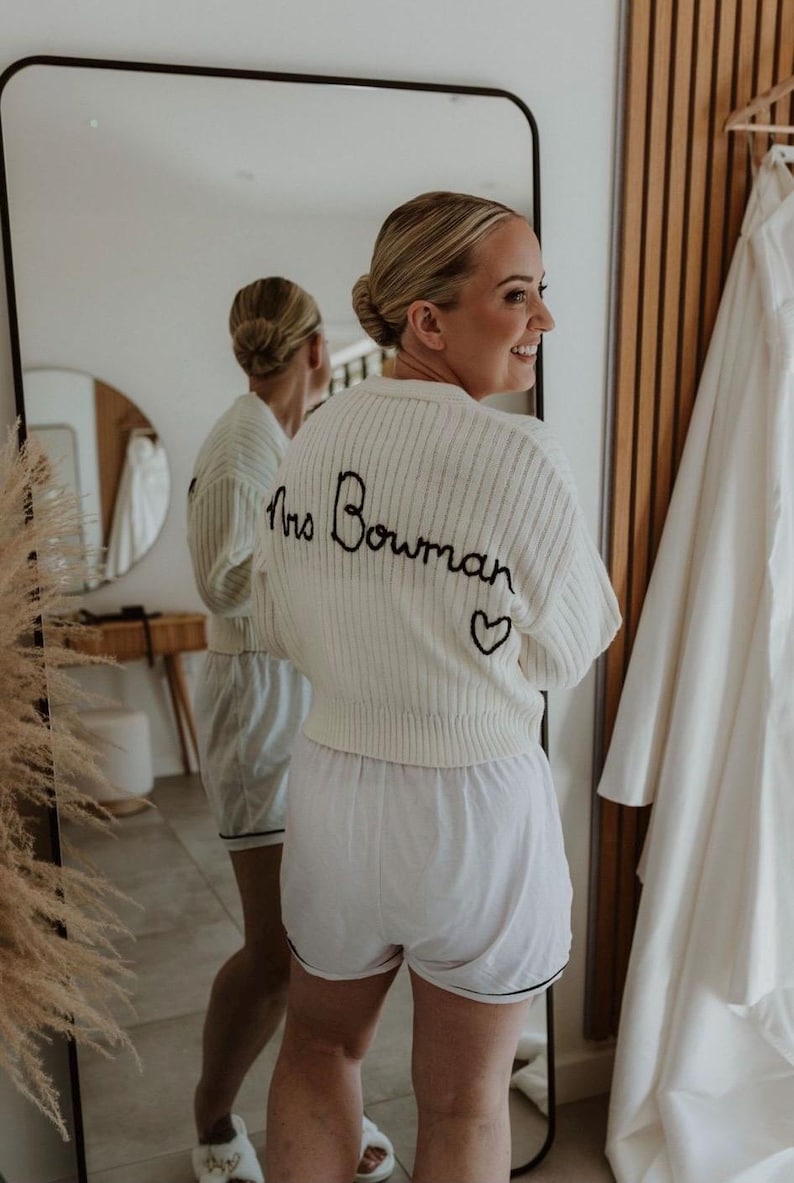 May include: A white knitted cardigan with the text "Mrs. Bowman" embroidered on the back. The cardigan has a small heart embroidered below the text.