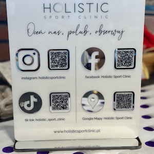 Puede incluir: Un letrero acrílico blanco para Holistic Sport Clinic, con el nombre de la clínica e iconos de redes sociales para Instagram, Facebook, TikTok y Google Maps, cada uno con un código QR. El letrero también incluye la dirección del sitio web.