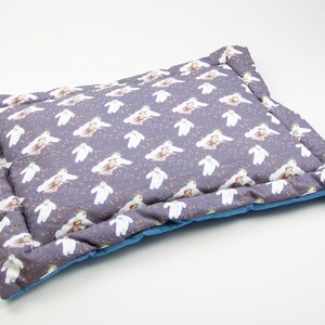 May include: A grey and blue rectangular pet bed with a white and grey patterned design featuring a repeating image of a bear wearing a hat. The bed has a blue border and a quilted design.