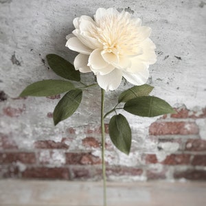 May include: A single white paper flower with green paper leaves on a stem. The flower is in focus and the background is blurred.