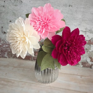 May include: Three paper flowers in a vase. The flowers are pink, white, and red. The vase is clear glass with a textured surface.