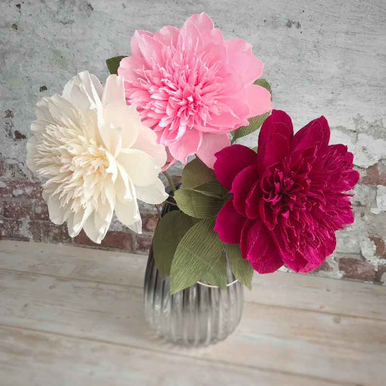 May include: Three paper flowers in a vase. The flowers are pink, white, and red. The vase is clear glass with a textured surface.