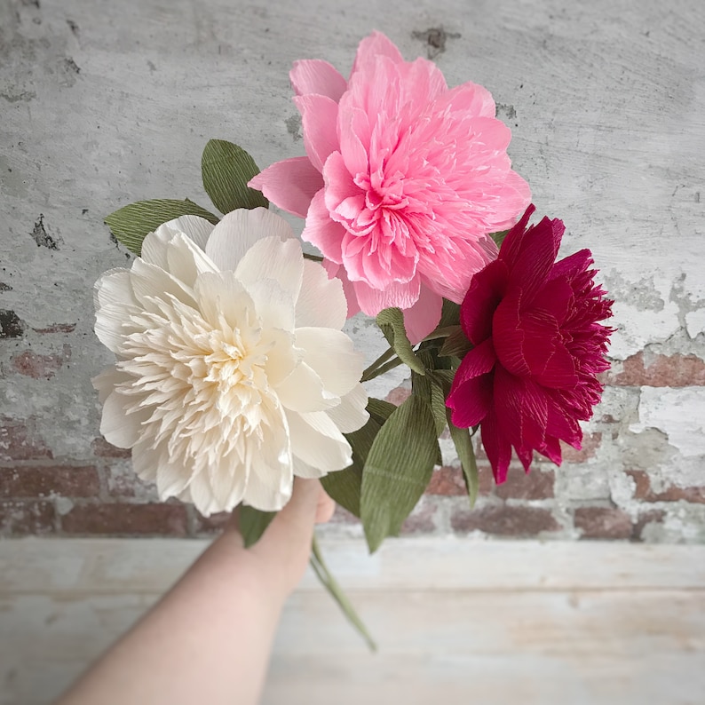 May include: Three paper peonies in pink, white, and red. The flowers are arranged in a bouquet and are held by a hand.