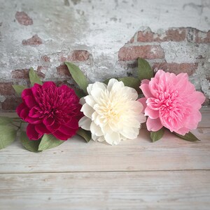 May include: Three paper peonies in different colors: pink, white, and burgundy. The flowers are arranged in a row on a light wood background.