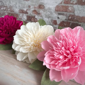 May include: Three large paper flowers in pink, white, and magenta. The flowers are made of crepe paper and have a ruffled texture.