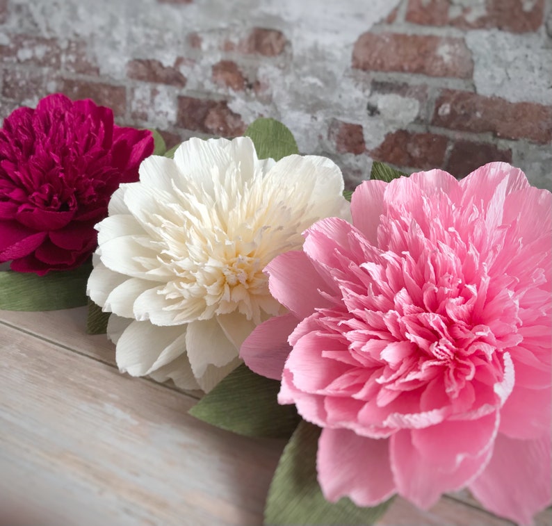 May include: Three large paper flowers in pink, white, and magenta. The flowers are made of crepe paper and have a ruffled texture.
