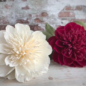 May include: Two large paper flowers, one white and one burgundy, with green paper leaves. The flowers are arranged on a white wooden surface.
