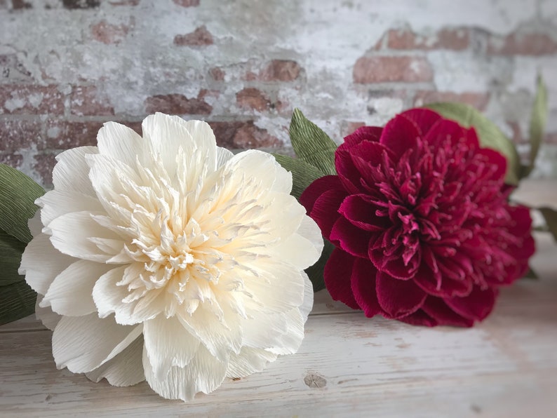 May include: Two large paper flowers, one white and one burgundy, with green paper leaves. The flowers are arranged on a white wooden surface.