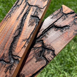 May include: Two wooden planks with a dark brown finish and intricate black fractal patterns. The wood grain is visible, and the planks are angled towards the viewer, showcasing the detailed burning technique. The background is blurred green grass.