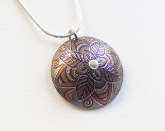 Könnte beinhalten: Eine silberne Kettenkette mit einem runden Anhänger mit einem violetten und goldenen Mandala-Design. Der Anhänger hat eine silberne Mitte.