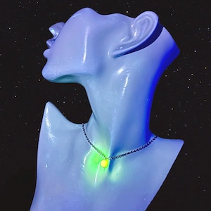 May include: A silver-toned metal chain necklace featuring a single, small, spherical pendant that emits a bright, glowing neon yellow light. The necklace is displayed on a smooth, light blue mannequin bust against a dark, starry background.