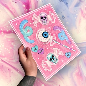 May include: A pink print featuring candy-themed illustrations. The design includes a lollipop with an eyeball, skull candies, and heart-shaped candies with the words "GO AWAY" and "NO". The print has a glittery effect.