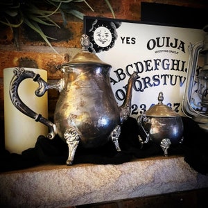 May include: A vintage silver tea set with a teapot and sugar bowl, displayed with a Ouija board and a candle. The teapot has ornate detailing and stands on three legs. The Ouija board features the words "YES" and "OUJIA" with the alphabet and numbers.