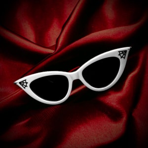 May include: White cat-eye sunglasses featuring black spiderweb graphics on the outer corners of the frames. The lenses are solid black, and the frames have a pointed, upswept shape at the temples, providing a distinct look for these eyewear accessories.