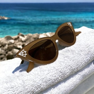 May include: Brown cat-eye sunglasses with dark lenses and a white spiderweb design on the outer corners. The sunglasses are resting on a white towel with a blue ocean background.