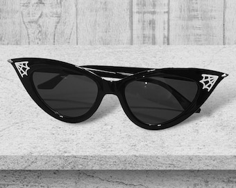 Puede incluir: Gafas de sol negras estilo ojo de gato con lentes oscuras y detalles de telaraña en tono plateado en las esquinas exteriores de la montura. Los marcos tienen una silueta afilada y puntiaguda con un acabado brillante.