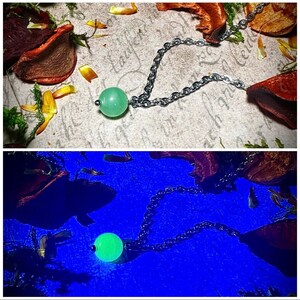 May include: A necklace featuring a light green, round gemstone pendant. The pendant is attached to a silver-colored chain. The background includes dried flowers and leaves, with a handwritten script visible in the background.