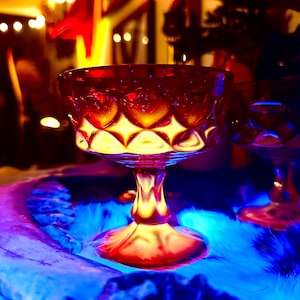 Puede incluir: Una copa de cristal de color rojo intenso con un patrón de diamantes texturizado en el cuenco. El tallo es corto y grueso, y termina en una base circular ancha. El cristal está iluminado por una luz cálida, creando reflejos brillantes.