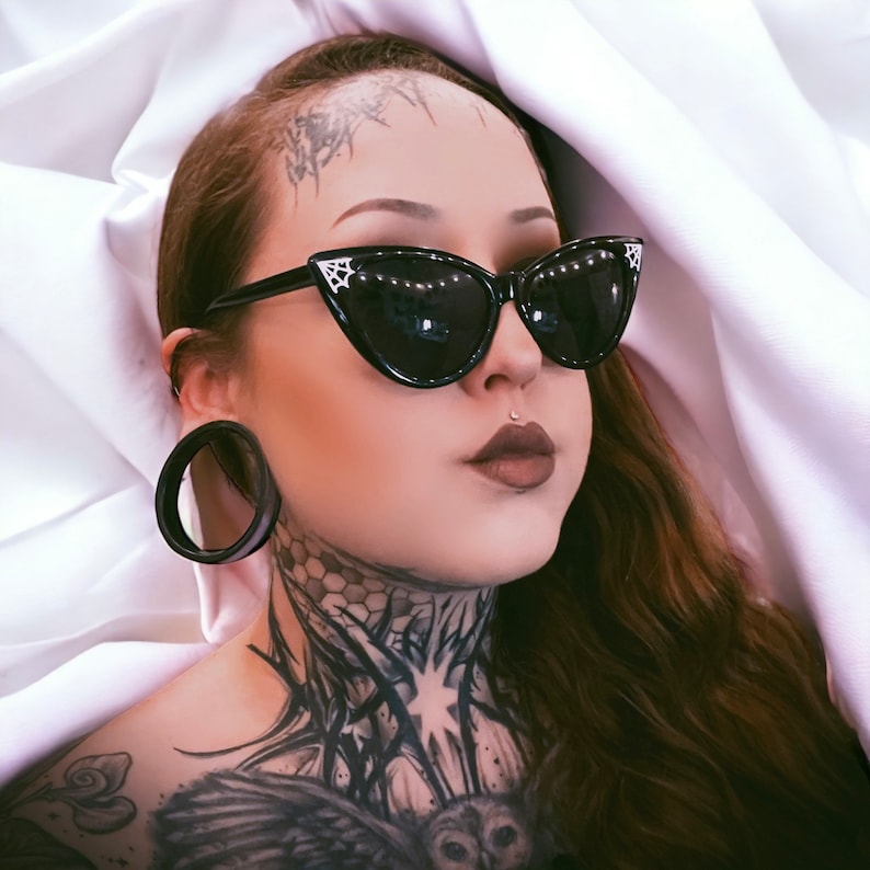 May include: A woman with black cat-eye sunglasses and a large black ear gauge. She has a detailed black and white tattoo on her neck and chest.
