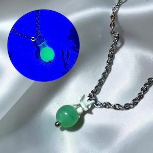 May include: A silver-toned chain necklace featuring a pendant with a green spherical bead attached to a white bone-like structure. An inset circular image shows the same pendant glowing with a bright green light against a dark blue background.