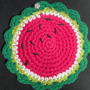 May include: A handmade, round crochet coaster in the shape of a watermelon slice. The coaster features a bright pink center with black seed details, surrounded by white, light green, and dark green edges. A small decorative charm is attached.