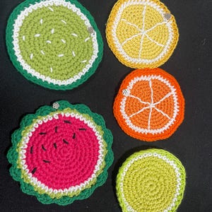 May include: A collection of five handmade crochet coasters in the shape of fruit slices. The coasters include a watermelon, lemon, lime, orange, and a green fruit slice. Each coaster has a white and green border.