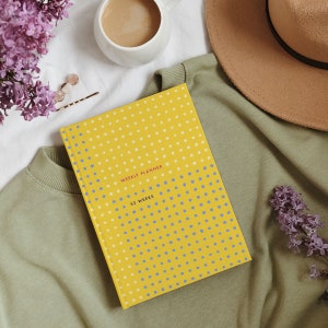 May include: A yellow weekly planner with a polka dot pattern in white and blue. The planner is open, displaying the text "WEEKLY PLANNER" and "52 WEEKS". The planner rests on a green sweatshirt, with a beige hat and a cup of coffee in the background.