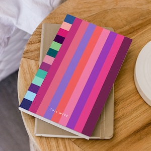May include: A colorful striped notebook with the text "THIS WEEK" on the cover. The notebook is closed and lying on a wooden surface.