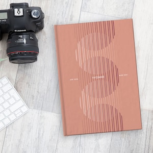 May include: A coral-colored journal with the name "YVONNE" and dates "APR 2026" and "MAY 2031" printed on the cover. The cover design features vertical lines and curved shapes. A camera, keyboard, and small plant are also visible.