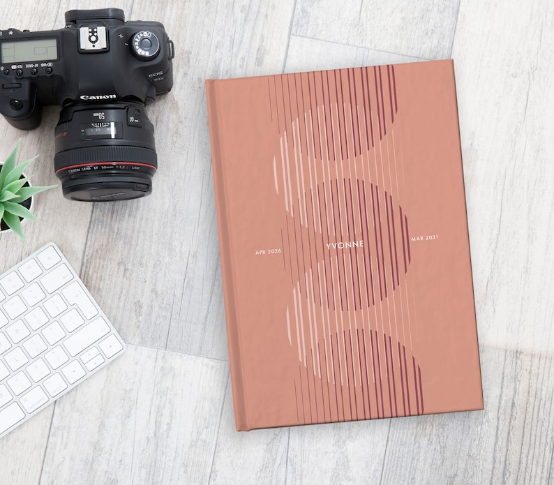 May include: A coral-colored journal with the name "YVONNE" and dates "APR 2026" and "MAY 2031" printed on the cover. The cover design features vertical lines and curved shapes. A camera, keyboard, and small plant are also visible.