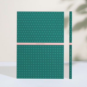 May include: A teal green notebook with a white polka dot pattern and a pink stripe across the bottom. The notebook has the name "Kamila Patel" printed on the stripe. The spine of the notebook is visible on the right side of the image and has the text "APR 2023 - MAR 2024" printed on it.