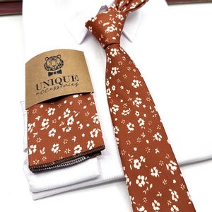 May include: A white dress shirt with a brown tie featuring a white floral pattern. The tie is knotted and the shirt is folded. A brown card with the text "UNIQUE accessories" and a tiger head logo is visible.