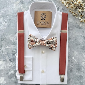 Peut inclure: Une chemise blanche avec un nœud papillon floral, des bretelles couleur rouille et une étiquette avec l'inscription "UNIQUE accessories". Le nœud papillon présente un fond crème avec des motifs floraux orange et marron. Les bretelles ont des attaches argentées.