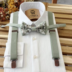 May include: A white textured dress shirt, complemented by sage green suspenders and a floral patterned bow tie. The bow tie displays a mix of white, grey, and black floral designs. The shirt has a small tag that reads "UNIQUE accessories".