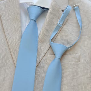 Può includere: Una cravatta azzurra e una cravatta a clip azzurra abbinata con cinturino regolabile e componenti in metallo argentato, mostrate su una camicia bianca con colletto e un blazer beige testurizzato con revers dentellati e taschino sul petto.