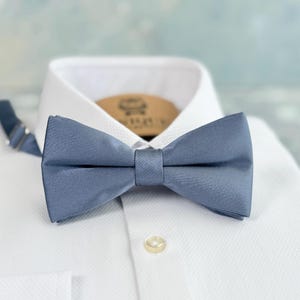 Könnte beinhalten: Eine staubblaue Fliege aus glattem Stoff ist auf einem weißen Hemd zentriert. Die Fliege ist sauber gebunden und hat eine klassische Schmetterlingsform. Das Hemd hat eine strukturierte Oberfläche und einen geknöpften Kragen.