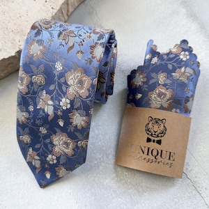Könnte beinhalten: Ein blaues Set aus Krawatte und Einstecktuch mit Blumenmuster. Krawatte und Einstecktuch zeigen ein sich wiederholendes Muster aus braunen und weißen Blumen auf blauem Hintergrund. Ein braunes Papieretikett mit dem Text "UNIQUE accessories" ist enthalten.