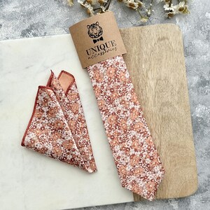 K&ouml;nnte beinhalten: Ein Set aus Krawatte und Einstecktuch mit floralem Muster. Die Krawatte ist in einer braunen Papph&uuml;lle mit dem Text "UNIQUE accessories" und einem Tiger-Logo verpackt. Das Einstecktuch ist gefaltet und hat ein passendes Blumenmuster. Das Muster umfasst Braunt&ouml;ne, Pfirsich und Wei&szlig;.