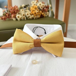 Può includere: Un papillon giallo senape con una fascia marrone effetto pelle. Il papillon è appoggiato su una camicia bianca con un motivo strutturato. È visibile un piccolo bottone rotondo.