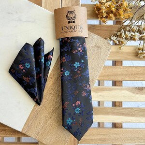 Op de afbeelding: Een zwarte stropdas en pochet set met een bloemenpatroon in blauw, roze en bruin. De stropdas wordt gepresenteerd met een bruin label met de tekst "UNIQUE accessories" en een tijgerlogo.