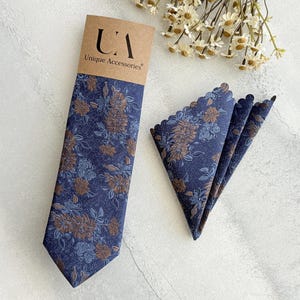 May include: A navy blue tie and pocket square set with a floral pattern in shades of brown and blue. The tie is displayed with its original packaging, which includes the text "Unique Accessories". The pocket square has a scalloped edge.