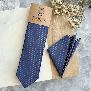 Peut inclure: Ensemble cravate et pochette de costume bleu marine à pois dorés. La cravate est emballée avec une carte marron avec l'inscription "UNIQUE accessories" et un logo de tigre. La pochette a un bord festonné.