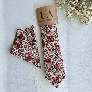 May include: A floral tie and pocket square set. The tie and pocket square feature a cream background with a pattern of red and pink roses and small flowers. The tie is attached to a brown cardboard tag with the text "UA Unique Accessories".