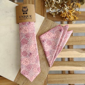 May include: A light pink tie and matching pocket square set with a floral pattern. The tie is displayed with a brown tag that reads "UNIQUE ACCESSORIES" and features a tiger logo. The floral pattern includes shades of pink and purple.