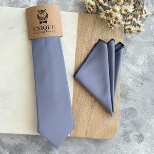 Op de afbeelding: Lichtblauwe stropdas en pochet set. De stropdas en pochet zijn gemaakt van een gladde stof. De set wordt gepresenteerd met een bruin label met de tekst "UNIQUE accessories" en een tijgerlogo. De pochet heeft een donkerblauwe rand.