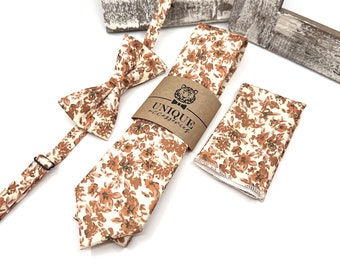 Op de afbeelding: Een set van drie bijpassende accessoires in een bloemenpatroon met oranje en bruine bloemen op een witte achtergrond. De set bevat een strik, een stropdas en een zakdoek. De stropdas heeft een bruin papieren etiket met de tekst "UNIQUE accessories".