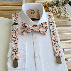 Puede incluir: Una camisa blanca con pajarita y tirantes florales. La pajarita y los tirantes presentan un fondo crema con un estampado de pequeñas flores naranjas, negras y grises. Los tirantes tienen clips metálicos.