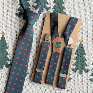 Puede incluir: Un conjunto de corbata y tirantes de color verde azulado con un estampado de lunares rojos. Los tirantes tienen clips metálicos plateados y un parche de cuero con un diseño verde y rojo. El conjunto se muestra sobre un soporte de cartón marrón.